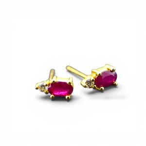 May include: A pair of gold stud earrings, each featuring an oval-cut vibrant pinkish-red gemstone set in a four-prong gold setting, accompanied by a single small, clear, round-cut accent stone positioned next to the primary oval gemstone.