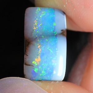 Könnte beinhalten: Ein rechteckiger Opal-Edelstein mit polierter Oberfläche. Der Opal zeigt ein lebendiges Farbspiel mit Blitzen in Blau, Grün und Orange. Der Stein hat eine weiße Basis mit braunen Einschlüssen an der Seite. Der Edelstein wird zwischen den Fingern gehalten.