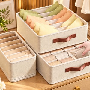 May include: Three beige fabric storage organizers with brown faux leather handles. The large square bin features multiple dividers holding pastel-colored items, while the two rectangular bins contain neatly folded beige garments in grid compartments.