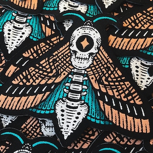 May include: Black embroidered patches featuring a moth design with a skull head and intricate wing patterns. The wings are detailed with orange and teal accents. The patches are likely for clothing or accessories.