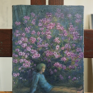 Op de afbeelding: Een schilderij van een persoon die voor een struik met roze bloemen zit. De persoon draagt een blauwe hoodie en heeft kort groen haar. De achtergrond is donkerblauw.