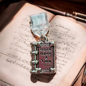 May include: A silver charm in the shape of a book with the words "Once Upon A Time..." on the cover. The charm is on a vintage book with handwritten text.
