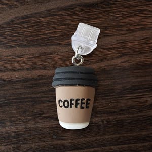 Peut inclure: Un charme en forme de tasse à café marron et noir avec le mot "COFFEE" imprimé sur le devant. Le charme est attaché à un clip en plastique transparent.