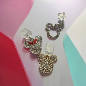 Puede incluir: Tres abalorios de cabeza de Mickey Mouse cubiertos de pedrería con clips de plástico transparente. Los abalorios son plateados, dorados y rojos con un lazo rojo.