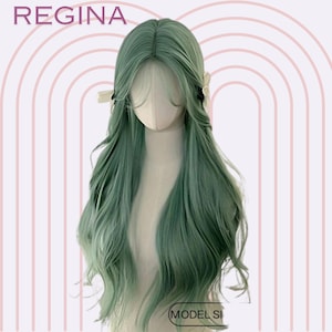 May include: A long, wavy wig in a muted green hue, styled on a mannequin head. The wig features a natural-looking part and flowing locks, with the word "REGINA" at the top. The bottom right corner of the image has the text "MODEL SI".