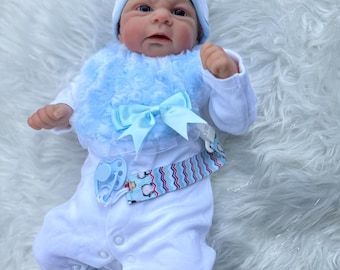May include: A realistic baby doll wearing a white onesie, blue bib, and matching hat. The bib has a bow, and the doll has a pacifier attached to a patterned strap. The doll is lying on a fluffy white surface.