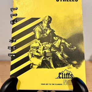 May include: A yellow and black spiral-bound book titled "Cliffs Notes on Shakespeare's Othello." The cover features a black and white illustration of a man and the Cliffs Notes logo. The book is displayed on a black stand.