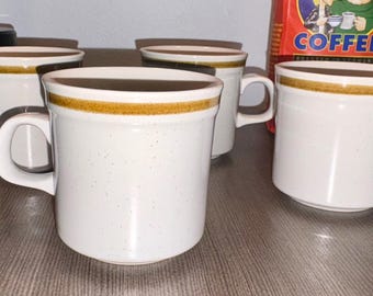 May include: Four white ceramic coffee mugs with a speckled finish and a brown band around the rim. Each mug has a large, curved handle. A bag of coffee is visible in the background.