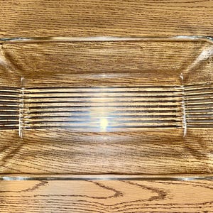 May include: A rectangular clear glass baking dish with ribbed detailing on the base and decorative handles. The dish is empty and sits on a wooden surface.