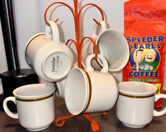 May include: A set of six off-white ceramic coffee mugs with brown trim, displayed on an orange metal mug tree. A bag of coffee with the text "Speeder & Earl's Coffee" is visible in the background. The mugs are labeled "Genuine Stoneware JAPAN".