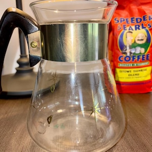 May include: A clear glass coffee carafe with a gold-colored metal band and a black handle. The carafe has a decorative starburst pattern and is next to a red bag of coffee with the text "Speeders & Earls Coffee".