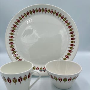 May include: A white dinnerware set featuring a large plate, a teacup, and a small bowl. Each piece has a repeating pattern of red, green, and gold decorative elements around the rim. The set is ideal for serving meals.