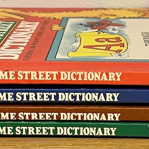 Puede incluir: Cuatro libros antiguos "Big Bird's Sesame Street Dictionary" apilados. Los libros son rojos, azules, marrones y verdes. Cada libro tiene el título impreso en el lomo. El libro superior tiene una ilustración de Big Bird.