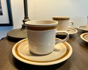 May include: Cream-colored ceramic coffee cups and saucers with a brown band around the rim. The cups have a handle and sit on matching saucers. The set is placed on a dark brown surface, with a lamp in the background.