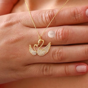 May include: A gold necklace with a pendant depicting two swans, each adorned with small, sparkling stones. The necklace is shown on a person's hand. The chain is delicate.