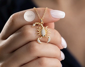 May include: A gold-toned scorpion pendant necklace featuring a delicate chain and a scorpion charm encrusted with small, clear, sparkling stones. The scorpion design includes detailed legs, pincers, and a curved tail. A circular logo reads CraftsByBaris.