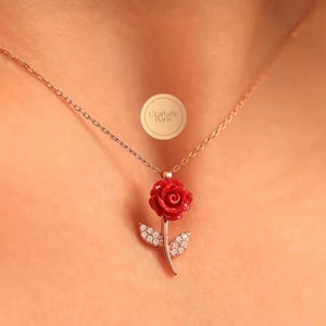 以下が含まれることがあります： 鮮やかな赤いバラの花が上部にあり、小さな透明な石が埋め込まれた2枚の葉を持つ茎が特徴のバラのペンダントネックレス。ペンダントは繊細なローズゴールドのチェーンから吊り下げられています。円形のロゴにはCraftsByBarisと記されています。
