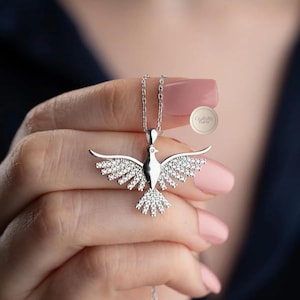 May include: A silver-colored pendant necklace featuring a dove with outstretched wings, encrusted with sparkling crystals. The dove pendant hangs from a delicate chain. The necklace is displayed against a blurred background.