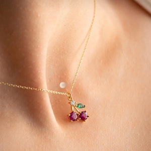 Op de afbeelding: Een goudkleurige ketting met een kersenhanger. De hanger heeft twee rode edelstenen, een groen blad en een kleine heldere edelsteen. De ketting wordt om de nek gedragen.