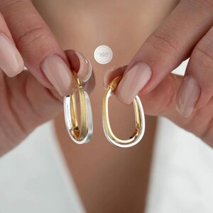 May include: Two gold and silver oval hoop earrings. One earring has a textured silver centre, while the other is solid gold. The earrings are held up against a white background. The earrings are a jewellery item.