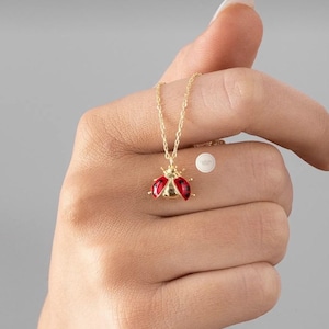 May include: A gold-colored necklace with a ladybug pendant. The ladybug has red and black enamel detailing. The necklace is held in a hand against a gray background. The ladybug pendant is approximately 0.5 inches in length.