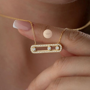 Könnte beinhalten: Eine goldfarbene Halskette mit einem horizontalen, länglichen ovalen Anhänger. Der Anhänger ist mit kleinen, klaren Pavé-Steinen besetzt und enthält drei größere, runde, klare Steine, die in einer Reihe in der Mitte gefasst sind.