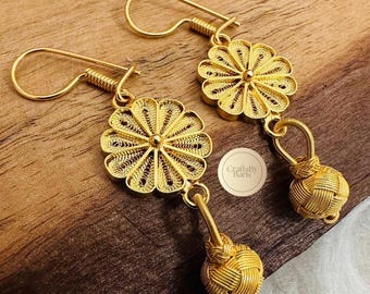 May include: A pair of gold-colored filigree earrings. Each earring features a flower-shaped top with a central detail and a woven knot pendant. The earrings have a hook closure. The image includes the text "CraftsByParis".