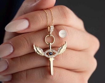 May include: Gold-coloured necklace with a pendant featuring an eye design, wings, and a sword-like element. The pendant is decorated with small, clear gemstones and a blue eye. The necklace is held by a person.