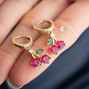 May include: Gold-tone hoop earrings with cherry pendants. Each pendant features three magenta-colored gemstones, a green leaf, and a small white gemstone. The earrings are designed to resemble cherries.