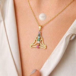 Peut inclure: Collier pendentif doré représentant une figure assise stylisée avec sept pierres précieuses colorées représentant les chakras. Le pendentif est suspendu à une chaîne délicate. Le collier est porté sur un chemisier crème.