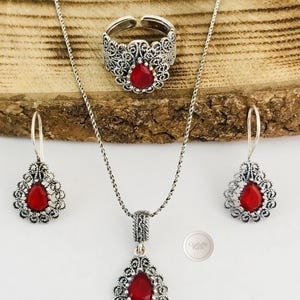 以下が含まれることがあります： ネックレス、指輪、イヤリングがセットになったシルバーのジュエリーセット。 各ピースには、精巧なシルバーの透かし細工のデザインに囲まれた赤い涙型の石があります。 セットは木製の表面に表示されます。