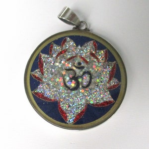May include: Circular pendant with a silver bail. The pendant features a blue background with a glittery lotus flower design in silver, red, and white. The Om symbol is in the center.