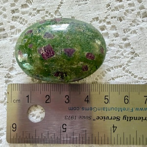 May include: An oval-shaped polished zoisite stone with deep green coloring and embedded magenta ruby crystals. A metal ruler is positioned directly beneath the stone, showing a length of approximately 2.25 inches for the gemstone.