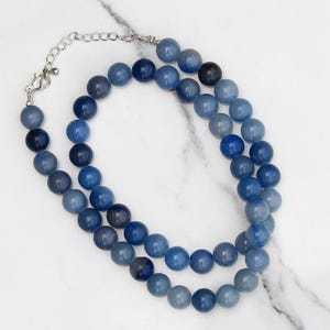 May include: A blue beaded necklace with a silver-colored chain and clasp. The necklace features two strands of round, blue beads in varying shades, creating a layered effect. The beads have a smooth, polished appearance.