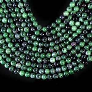 May include: A strand of green, black, and pink gemstone beads. The beads are round and have a speckled appearance.