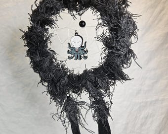May include: A handmade dreamcatcher with a dark gray, textured wreath design. The center features a white web with a cartoon octopus in a suit. Two black feathers hang below, suspended by a chain.