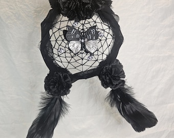 May include: A black dreamcatcher with a butterfly design in the center. The dreamcatcher features black feathers, pom-poms, and a webbed interior. The butterfly has silver and black details. The dreamcatcher is hanging against a white background.
