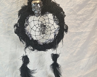 May include: A black and silver Halloween dreamcatcher with a skull and spider design. The dreamcatcher features a black web, black feathers, and a silver skull at the top. The design includes small skull accents around the web.