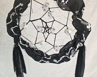 May include: A Halloween-themed dreamcatcher with a black and white skeleton print fabric wrapped around the hoop. The dreamcatcher features white string webbing, small ghost-shaped charms with red accents, and black feathers.