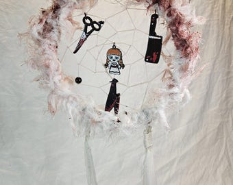 May include: A handmade dreamcatcher with a white web and a fluffy, textured outer ring with maroon accents. It features small charms of scissors, a cleaver, a doll, and a knife, all with a horror theme. White feathers hang from the bottom.