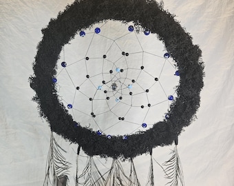 May include: A handcrafted dreamcatcher with a black moss-covered hoop. The web is strung with black beads and blue glass beads. Peacock feathers hang from the bottom, creating a bohemian aesthetic. The dreamcatcher is suspended by a black ribbon.