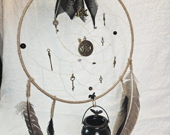 May include: A handcrafted dreamcatcher with a woven web, adorned with small antique keys, feathers, and a black cauldron. A large black bow with a key and moon charm tops the dreamcatcher. The dreamcatcher is designed with a rustic, witchy aesthetic.