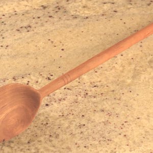 Könnte beinhalten: Ein Holzlöffel mit langem Stiel und abgerundeter Schüssel. Der Löffel ist aus hellbraunem Holz gefertigt und hat eine glatte Oberfläche. Der Griff hat ein paar dekorative Rillen in der Nähe der Oberseite.