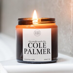 May include: A brown glass candle jar with a black label that reads "this candle smells like COLE PALMER" and "scented soy candle 180ml / 6oz". The candle is lit and the flame is visible.
