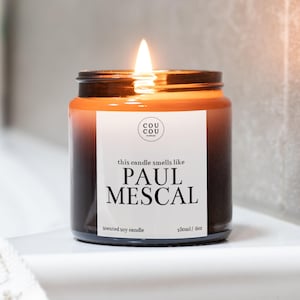 May include: A brown glass candle jar with a black label that reads "this candle smells like PAUL MESCAL" and "scented soy candle 180ml / 6oz". The candle is lit and the flame is visible.