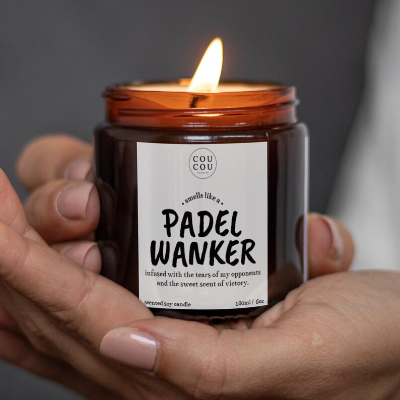 May include: A lit, brown glass jar candle with a white label that reads "PADEL WANKER". The candle is labeled "scented soy candle" and has a 6 oz volume. The candle is held in a person's hands.
