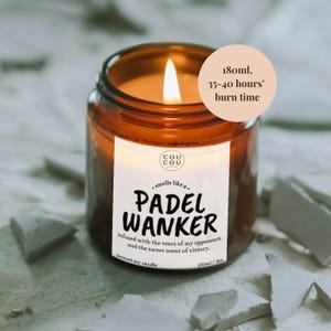 May include: A lit scented soy candle in a brown glass jar. The label reads "PADEL WANKER" with text about the scent. The candle has a burn time of 35-40 hours and contains 6 oz of wax.