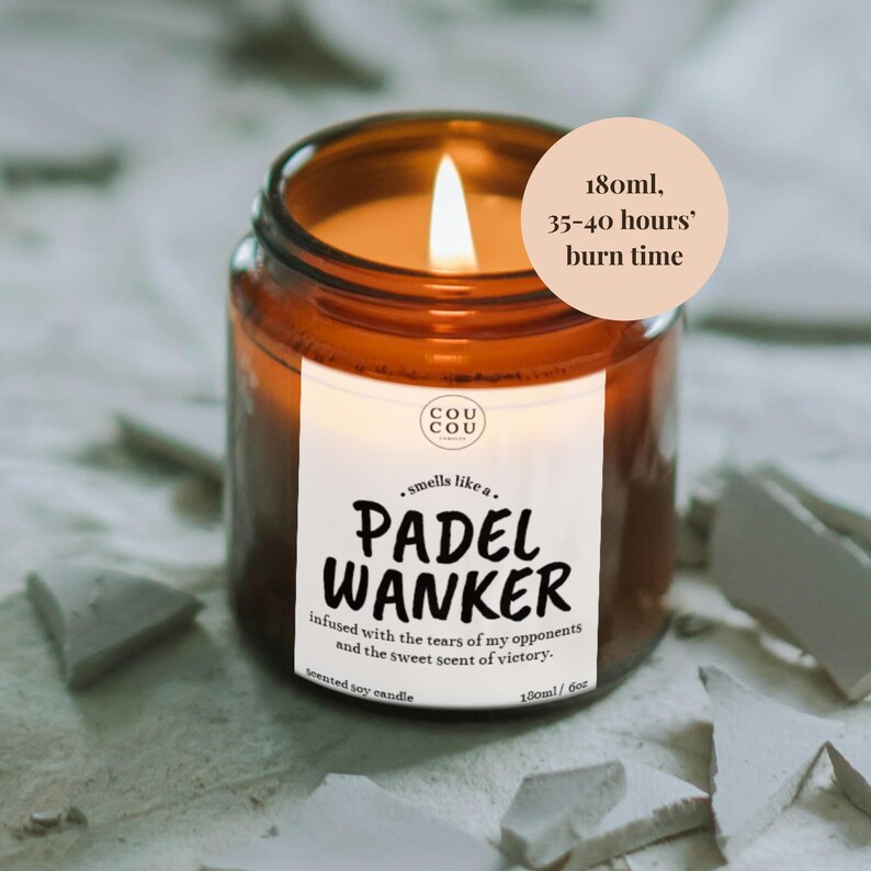 May include: A lit scented soy candle in a brown glass jar. The label reads "PADEL WANKER" with text about the scent. The candle has a burn time of 35-40 hours and contains 6 oz of wax.