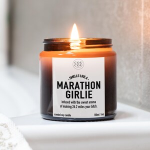 May include: A lit scented soy candle in a dark amber glass jar with a white label. The label features the text "COU COU", "SMELLS LIKE A MARATHON GIRLIE", and "infused with the sweet aroma of making 26.2 miles your bitch." It is marked 180ml / 6oz.