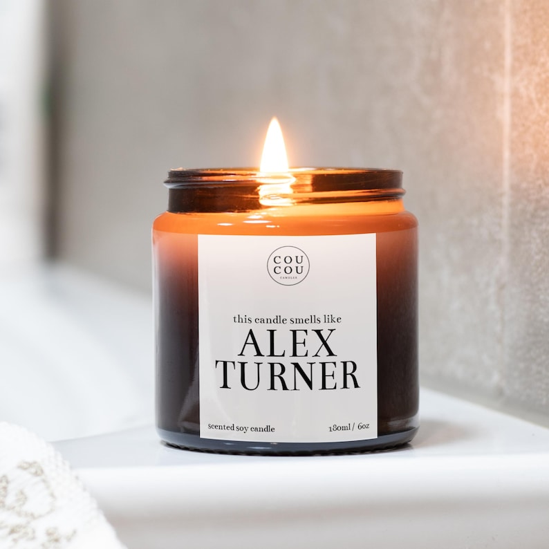 May include: A brown glass candle with a black label that reads "this candle smells like ALEX TURNER" and "scented soy candle 180ml / 6oz". The candle is lit and the flame is visible.